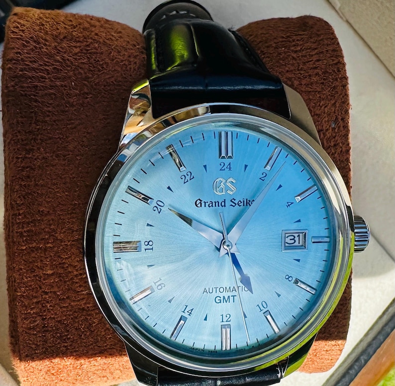 May include: A Grand Seiko automatic GMT watch with a light blue sunray dial, silver-tone indices, and a date window at the three o'clock position. The timepiece features a polished stainless steel case and a black leather strap with a buckle.