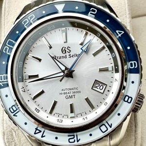 May include: A Grand Seiko automatic watch with a silver dial and a blue and white bezel. The watch has a date display and GMT function. The watch has a stainless steel band. The watch face displays the text "AUTOMATIC HI-BEAT 36000 GMT".