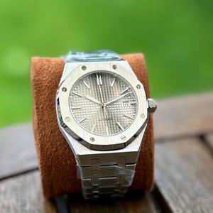 May include: A silver-toned, automatic wristwatch with a textured dial and a date display. The watch features a stainless steel bracelet and a distinctive octagonal bezel. The watch is displayed on a brown cushion against a blurred green background.