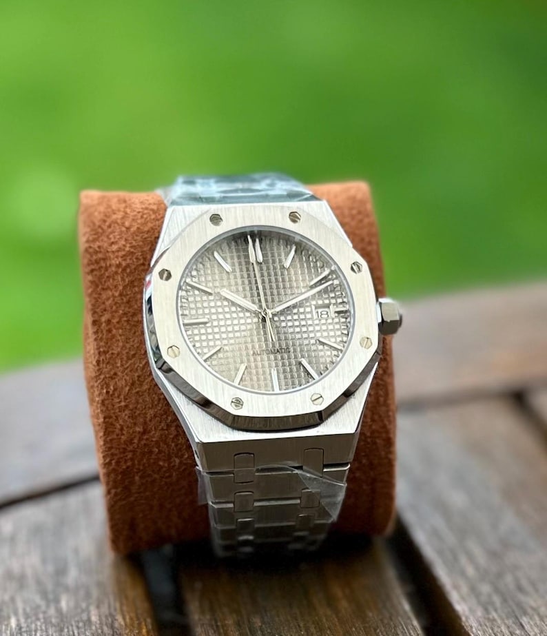 May include: A silver-toned, automatic wristwatch with a textured dial and a date display. The watch features a stainless steel bracelet and a distinctive octagonal bezel. The watch is displayed on a brown cushion against a blurred green background.