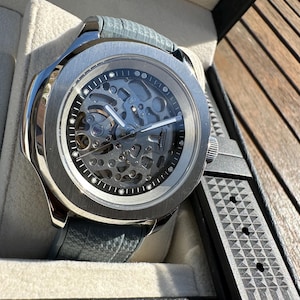 Peut inclure: Une montre squelette argent&eacute;e avec un bracelet en cuir gris et un bracelet en caoutchouc noir. Le cadran r&eacute;v&egrave;le les m&eacute;canismes internes complexes. La montre est pr&eacute;sent&eacute;e dans un coffret noir avec un int&eacute;rieur gris clair.