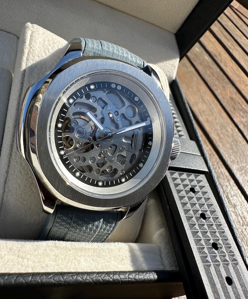 Peut inclure: Une montre squelette argent&eacute;e avec un bracelet en cuir gris et un bracelet en caoutchouc noir. Le cadran r&eacute;v&egrave;le les m&eacute;canismes internes complexes. La montre est pr&eacute;sent&eacute;e dans un coffret noir avec un int&eacute;rieur gris clair.