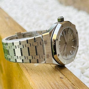 May include: A silver-toned wristwatch with a textured dial and a matching bracelet. The watch is displayed on a wooden surface, with a blurred green lawn and white pebbles in the background. The watch has a round face and a crown.