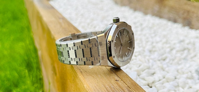 May include: A silver-toned wristwatch with a textured dial and a matching bracelet. The watch is displayed on a wooden surface, with a blurred green lawn and white pebbles in the background. The watch has a round face and a crown.