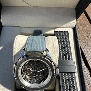 Peut inclure: Une montre squelette argent&eacute;e avec un bracelet gris et un bracelet noir de rechange, pr&eacute;sent&eacute;e dans un coffret noir avec un int&eacute;rieur gris clair. Le cadran r&eacute;v&egrave;le les m&eacute;canismes internes.