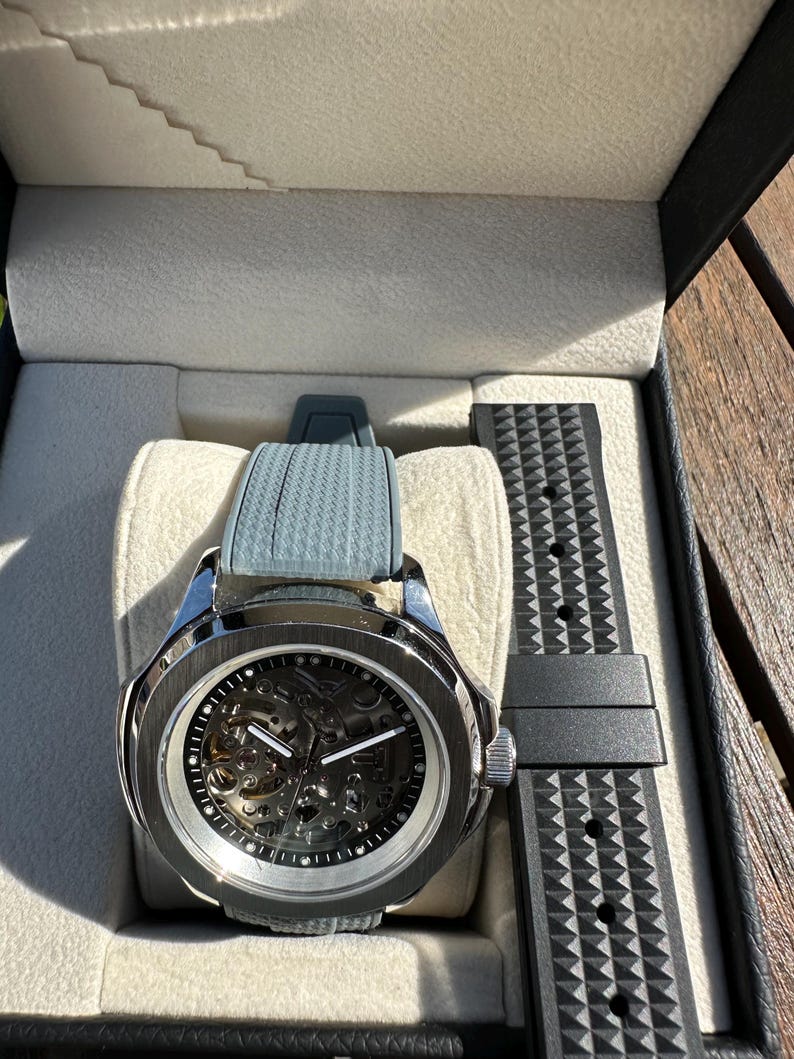 Peut inclure: Une montre squelette argent&eacute;e avec un bracelet gris et un bracelet noir de rechange, pr&eacute;sent&eacute;e dans un coffret noir avec un int&eacute;rieur gris clair. Le cadran r&eacute;v&egrave;le les m&eacute;canismes internes.