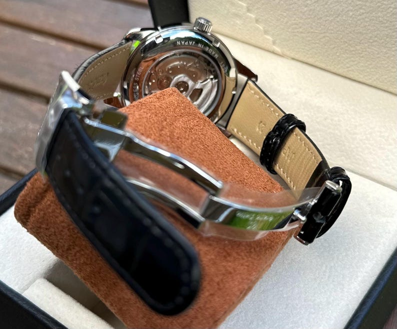 May include: A stainless steel wristwatch with a black leather strap and a deployment clasp. The watch features an exhibition case back revealing the internal movement, with the text MADE IN JAPAN and STAINLESS STEEL engraved on the metal casing.