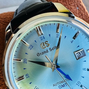 May include: A Grand Seiko automatic GMT watch with a light blue sunray dial, silver-tone indices, and a date window at the three o'clock position. The watch features a polished silver-tone case and a black leather strap with a visible gold GS logo.