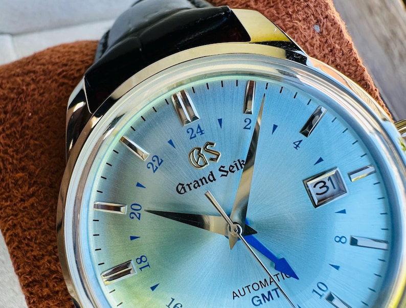 May include: A Grand Seiko automatic GMT watch with a light blue sunray dial, silver-tone indices, and a date window at the three o'clock position. The watch features a polished silver-tone case and a black leather strap with a visible gold GS logo.