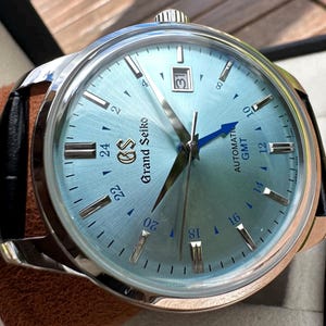 May include: A Grand Seiko automatic GMT watch with a light blue sunray dial, silver-tone indices, and a date window at the three o'clock position. The watch features a blue GMT hand, a stainless steel case, and a black leather strap.