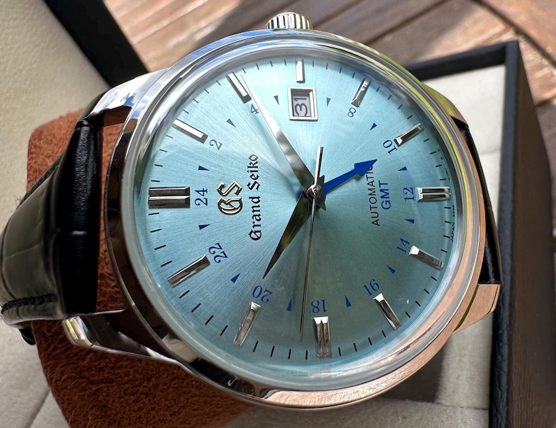 May include: A Grand Seiko automatic GMT watch with a light blue sunray dial, silver-tone indices, and a date window at the three o'clock position. The watch features a blue GMT hand, a stainless steel case, and a black leather strap.