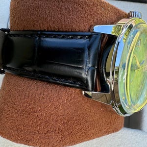 May include: A wristwatch featuring a polished silver-tone metal case and a black leather strap with a visible grain texture. The watch face displays a bright lime green dial with thin metallic hands and a small silver crown on the side.