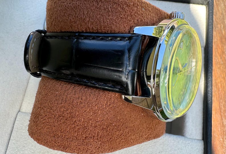May include: A wristwatch featuring a polished silver-tone metal case and a black leather strap with a visible grain texture. The watch face displays a bright lime green dial with thin metallic hands and a small silver crown on the side.