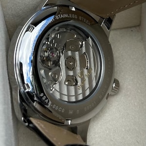 May include: A stainless steel wristwatch with an exhibition case back revealing the internal mechanical movement. The metal casing is engraved with Water Resistant, Made in Japan, and the model number 9SA5-00C0. A black leather strap is attached.
