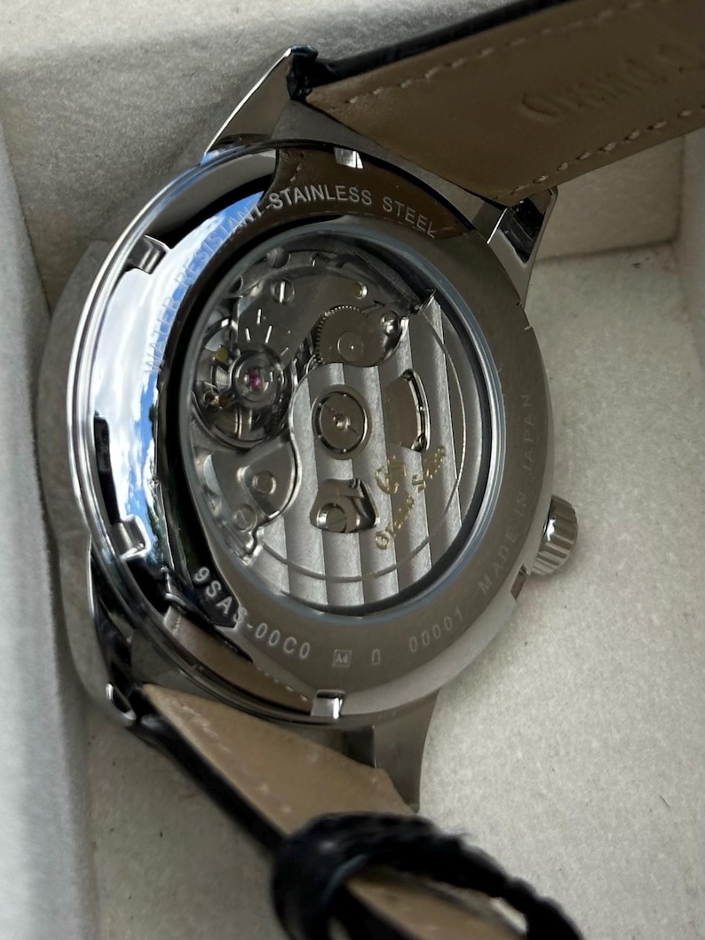 May include: A stainless steel wristwatch with an exhibition case back revealing the internal mechanical movement. The metal casing is engraved with Water Resistant, Made in Japan, and the model number 9SA5-00C0. A black leather strap is attached.