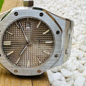 May include: A silver-toned wristwatch with a textured, brown face and silver hands. The watch has a stainless steel band and a date display at the 3 o'clock position. The watch is on a wooden surface.