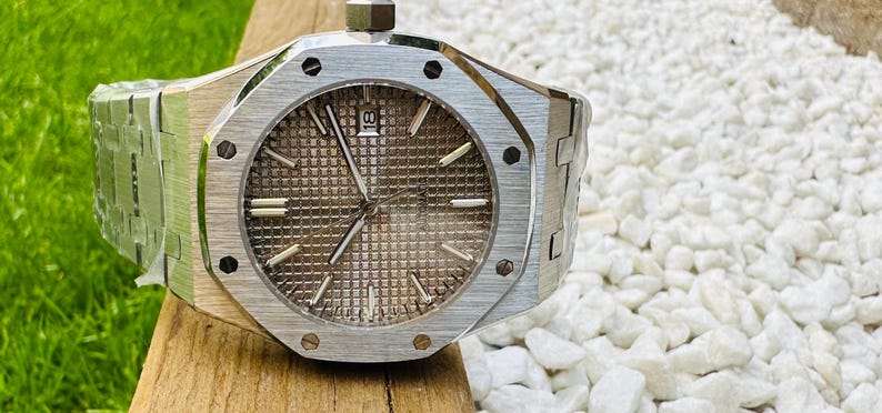 May include: A silver-toned wristwatch with a textured, brown face and silver hands. The watch has a stainless steel band and a date display at the 3 o'clock position. The watch is on a wooden surface.