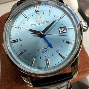 May include: A Grand Seiko automatic GMT watch with a light blue sunray dial, silver-tone hour markers, and a date window at the three o'clock position. The watch features a bright blue GMT hand, a stainless steel case, and a black leather strap.