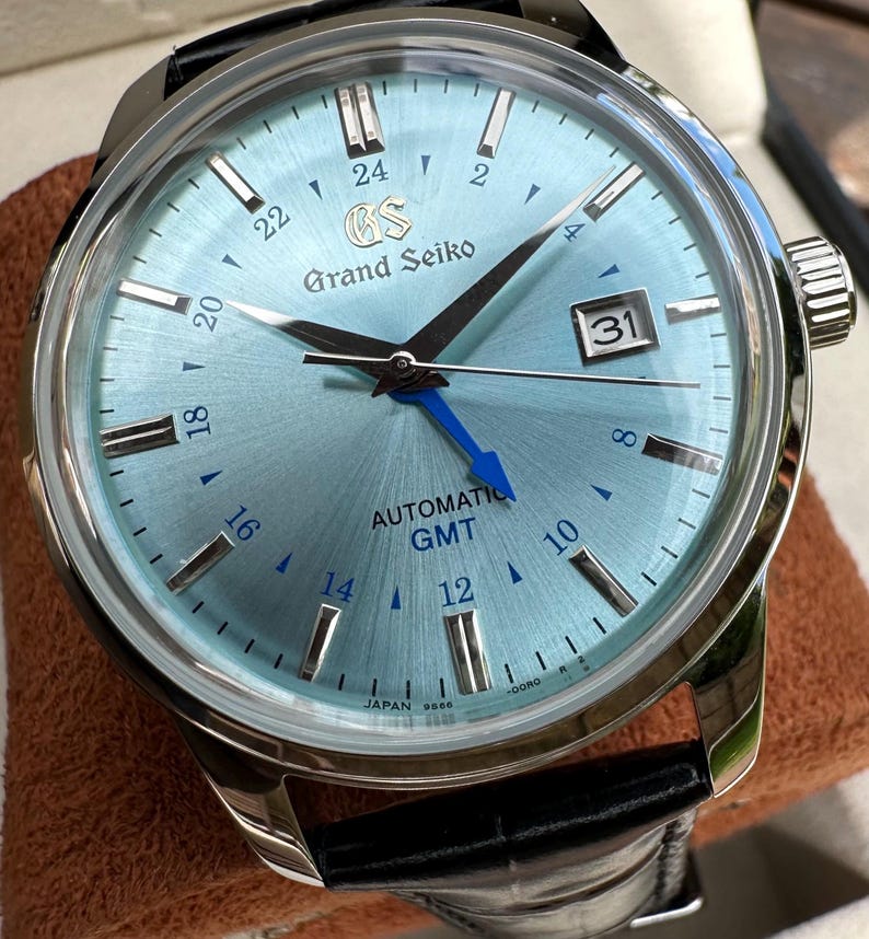 May include: A Grand Seiko automatic GMT watch with a light blue sunray dial, silver-tone hour markers, and a date window at the three o'clock position. The watch features a bright blue GMT hand, a stainless steel case, and a black leather strap.