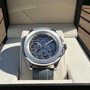 Peut inclure: Une montre squelette argent&eacute;e avec un bracelet en cuir bleu, pr&eacute;sent&eacute;e dans un coffret noir avec un int&eacute;rieur cr&egrave;me. Le cadran r&eacute;v&egrave;le le m&eacute;canisme interne de la montre.