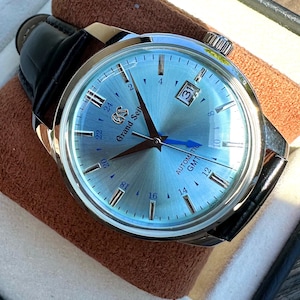 May include: A Grand Seiko automatic GMT watch with a light blue sunray dial, silver-tone indices, and a date window at the three o'clock position. The watch features a blue GMT hand, silver hour and minute hands, and a black leather strap.