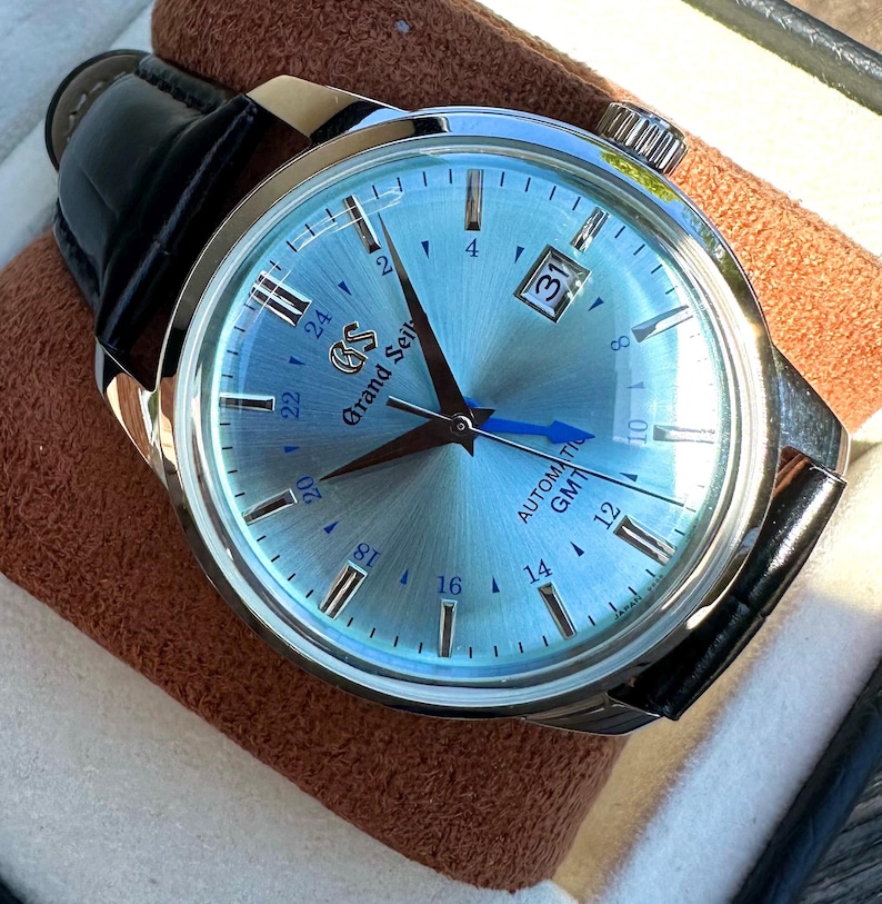 May include: A Grand Seiko automatic GMT watch with a light blue sunray dial, silver-tone indices, and a date window at the three o'clock position. The watch features a blue GMT hand, silver hour and minute hands, and a black leather strap.