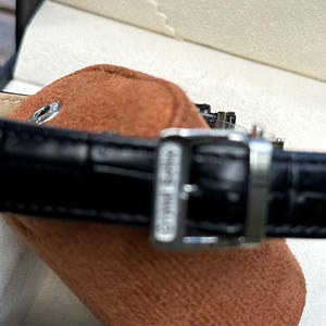 May include: A wristwatch featuring a polished silver-tone metal case and a black leather strap with a crocodile-embossed texture. The strap includes a silver-tone metal buckle with the text Grand Seiko engraved on the side of the buckle hardware.