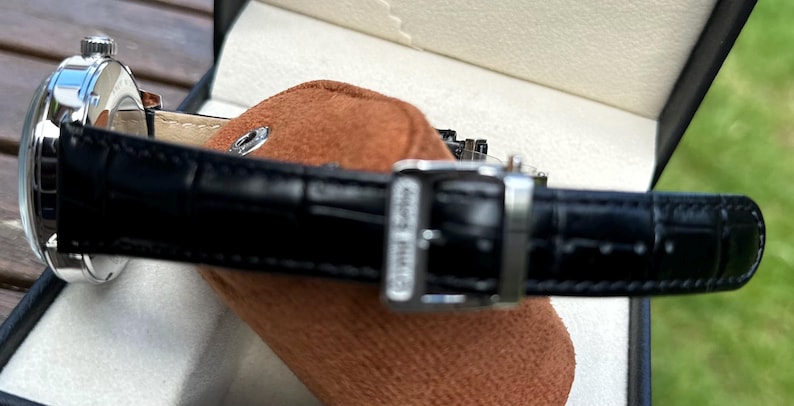 May include: A wristwatch featuring a polished silver-tone metal case and a black leather strap with a crocodile-embossed texture. The strap includes a silver-tone metal buckle with the text Grand Seiko engraved on the side of the buckle hardware.