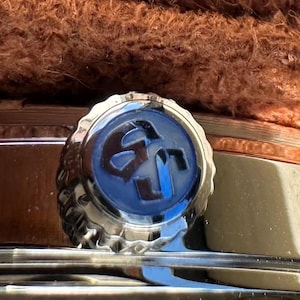May include: A silver-toned watch crown featuring a circular blue inlay with a dark embossed logo. The crown is positioned on the side of a metallic watch case, partially obscured by a textured brown material resting along the top edge of the watch.