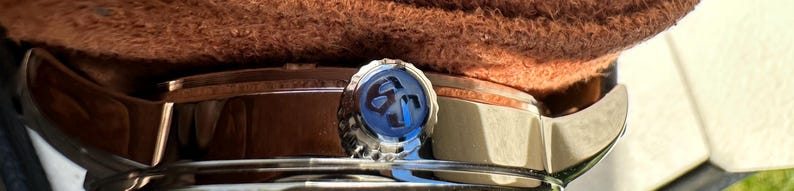 May include: A silver-toned watch crown featuring a circular blue inlay with a dark embossed logo. The crown is positioned on the side of a metallic watch case, partially obscured by a textured brown material resting along the top edge of the watch.