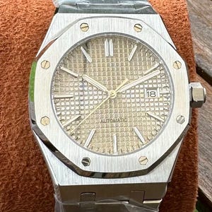 May include: Silver-toned wristwatch with a gold-colored face and a date display. The watch has a textured dial, luminous hands, and a stainless steel band. The watch is labeled "AUTOMATIC".