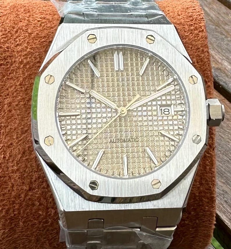May include: Silver-toned wristwatch with a gold-colored face and a date display. The watch has a textured dial, luminous hands, and a stainless steel band. The watch is labeled "AUTOMATIC".