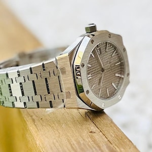 May include: A silver-toned wristwatch with a textured gray face and a date display. The watch features a stainless steel band with a repeating link pattern. The watch is set on a wooden surface.