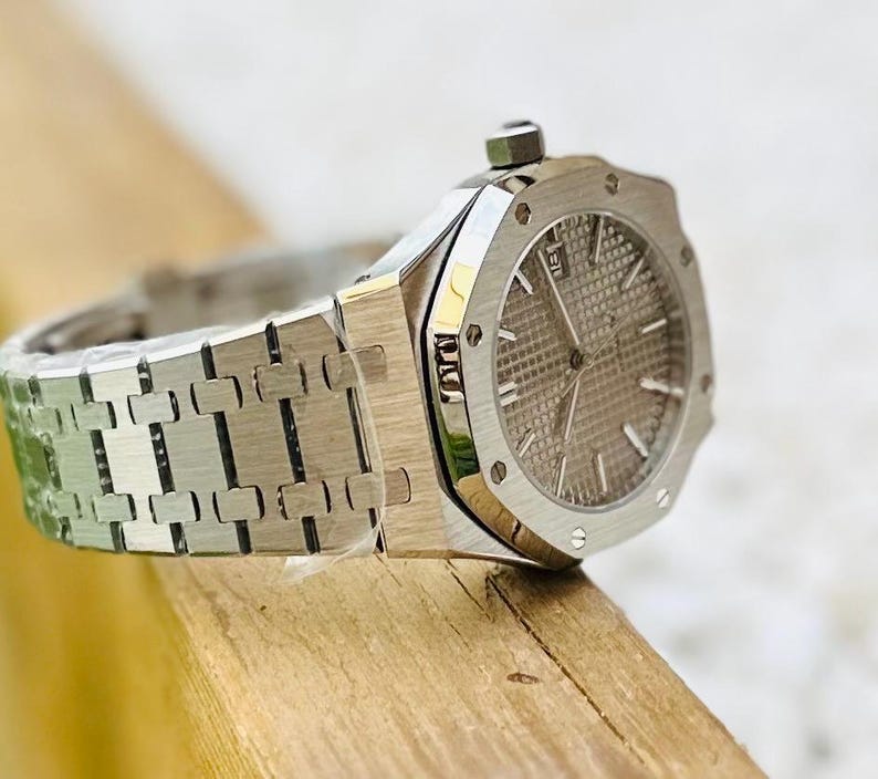 May include: A silver-toned wristwatch with a textured gray face and a date display. The watch features a stainless steel band with a repeating link pattern. The watch is set on a wooden surface.