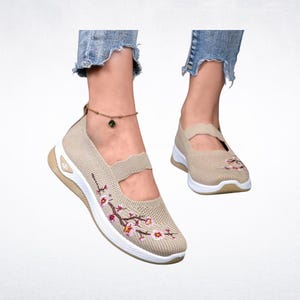 Pu&ograve; includere: Scarpe Mary Jane in maglia beige con suola bianca e tacco piccolo. Le scarpe presentano un ricamo floreale rosa e marrone. Le scarpe hanno un cinturino sulla parte superiore del piede.