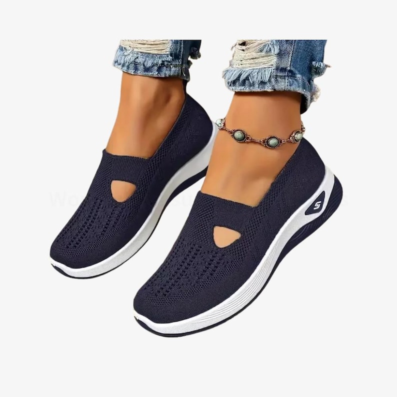 May include: Navy blue slip-on trainers with a white sole and a small cutout detail on the top. The shoes have a breathable knit upper and a sporty design. The trainers are shown on a person wearing blue jeans.