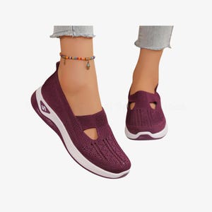 May include: Plum-coloured slip-on trainers with a white sole. The shoes have a breathable knit upper and a unique cut-out design on the top. The trainers are shown on a person's feet.