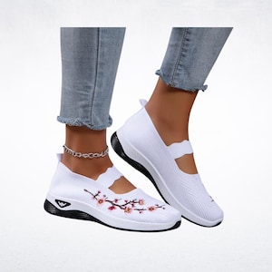 Pu&ograve; includere: Scarpe bianche slip-on con ricamo floreale. Le scarpe hanno suola nera e tomaia bianca con cinturino decorativo. Il design floreale presenta fiori rosa e arancioni con foglie verdi.
