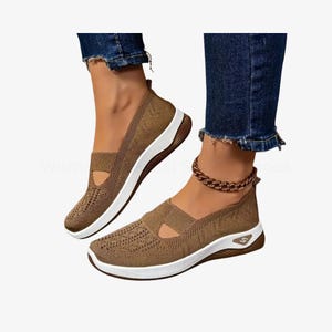 May include: Brown slip-on trainers with a breathable knit upper and white soles. The shoes have a cut-out detail on the top and a small logo on the side. The trainers are paired with blue jeans.