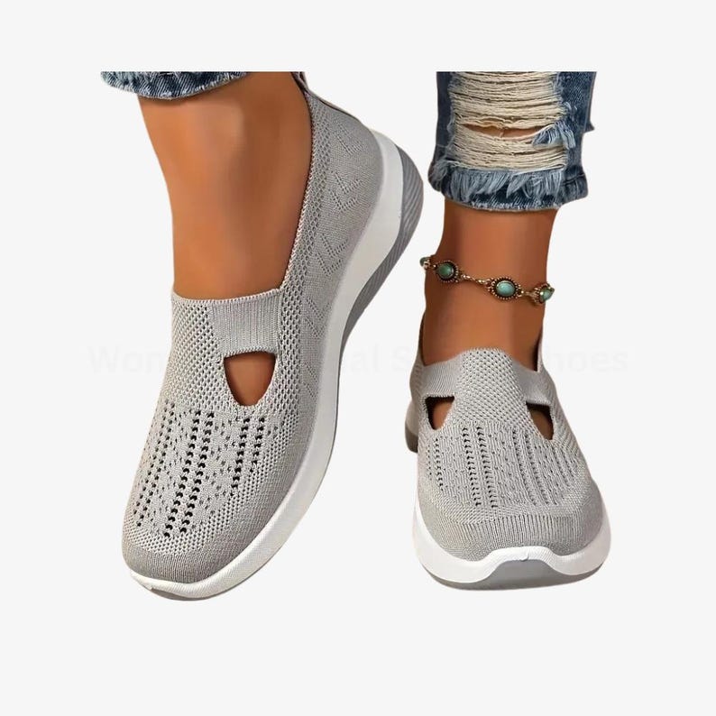 May include: Grey slip-on trainers with a breathable knit upper and a white sole. The shoes feature a distinctive cut-out design on the top, designed for comfort and style. The trainers are shown on a person's feet.
