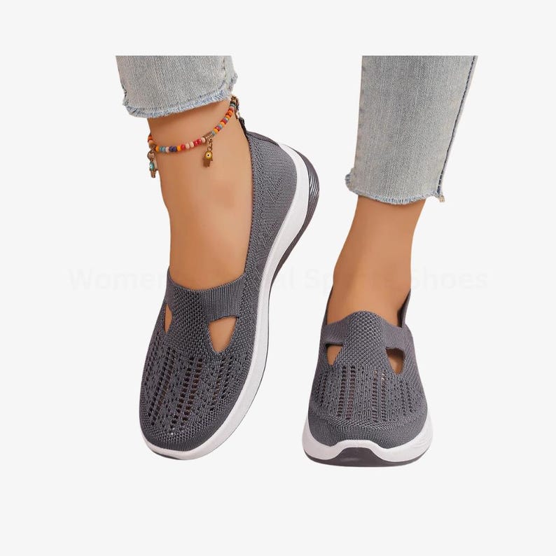 May include: Grey slip-on shoes with a breathable knit upper and a white sole. The shoes have a unique cut-out design on the top. The shoes are shown on a person's feet. These shoes are designed for comfort and style.