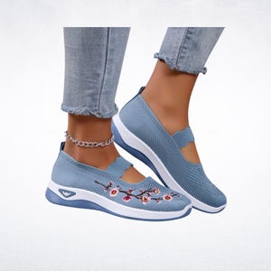 Pu&ograve; includere: Sneakers slip-on azzurre con suole bianche e ricami floreali. Le scarpe hanno una tomaia in maglia traspirante e un bordo smerlato. Le sneakers sono mostrate con jeans azzurri.