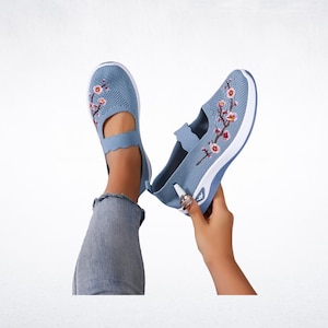 Pu&ograve; includere: Scarpe slip-on in rete blu chiaro con ricami floreali. Le scarpe hanno una suola bianca e un cinturino smerlato. Una scarpa &egrave; su un piede e l'altra &egrave; tenuta in mano.