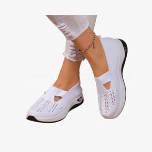 May include: White slip-on trainers with a breathable knit upper and a black sole. The shoes have a unique cut-out design on the top and a cushioned sole for comfort. The trainers are designed for casual wear.