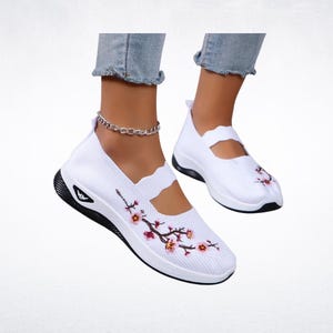 Pu&ograve; includere: Sneakers bianche slip-on con suola nera e ricamo floreale. Le scarpe hanno una tomaia in maglia traspirante e un cinturino decorativo. Le sneakers sono mostrate su una persona che indossa jeans blu.