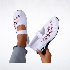 Pu&ograve; includere: Scarpe bianche slip-on con ricamo floreale. Le scarpe hanno una tomaia in maglia traspirante, un cinturino smerlato e una suola bianca e nera. Il ricamo floreale presenta fiori rosa e rossi con steli verdi.