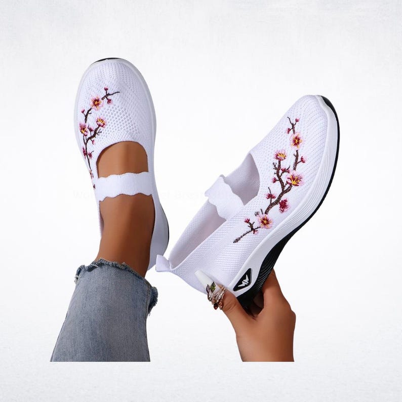 Pu&ograve; includere: Scarpe bianche slip-on con ricamo floreale. Le scarpe hanno una tomaia in maglia traspirante, un cinturino smerlato e una suola bianca e nera. Il ricamo floreale presenta fiori rosa e rossi con steli verdi.