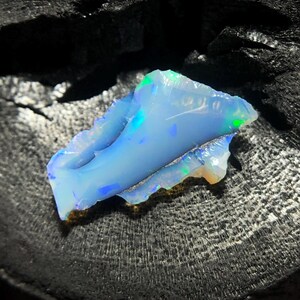 Könnte beinhalten: Ein roher, ungeschliffener Opal-Edelstein mit hellblauer Grundfarbe und schillernden grünen und blauen Blitzen. Der Opal hat eine raue, unebene Textur und ist vor einem dunklen, strukturierten Hintergrund platziert.