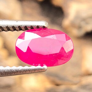 Könnte beinhalten: Ein facettierter, oval geschliffener Rubin, gehalten von einer silbernen Pinzette. Der Rubin hat eine leuchtend rosa Farbe und viele Facetten, die das Licht reflektieren. Der Hintergrund ist ein verschwommenes Holzmaserungsmuster.