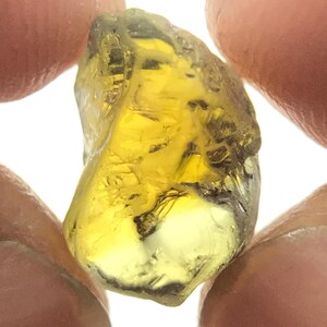 May include: A rough-cut, translucent yellow gemstone held between two fingers. The gem has a natural, irregular shape with a slightly darker hue in some areas. The stone appears to be a raw, unpolished mineral.